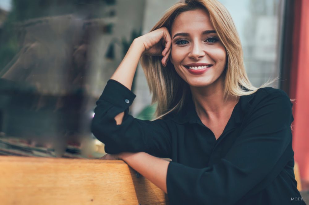 A young blonde woman wearing a black blouse rests her head on her hand and smiles at the camera