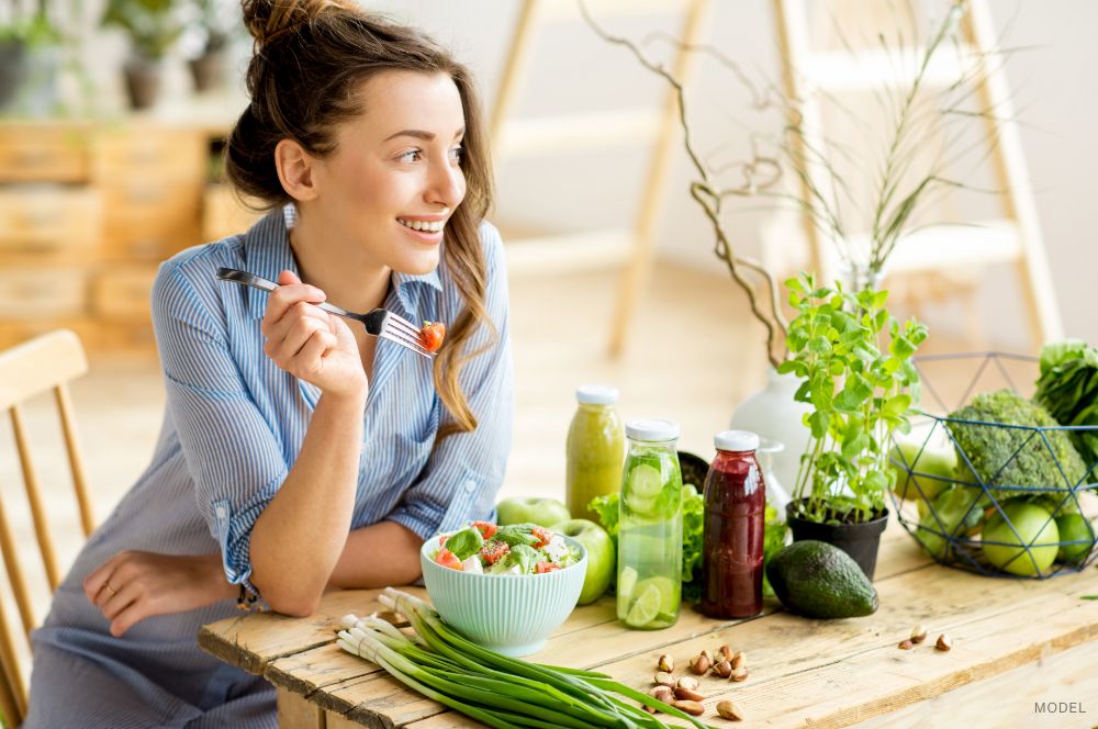 A young woman in a casual blue dress and with her hair in a messy bun smiles while seated at a wooden table eating a salad